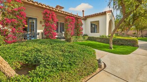 A house with a tiled roof, a patio with outdoor furniture, and a well-maintained garden with hedges and flowering plants. - LV021 A Single Story Studio Near the Main Pools (La Quinta)