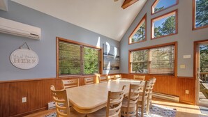 Un comedor con una mesa y sillas de madera, ventanas amplias con persianas y un aire acondicionado de pared.
