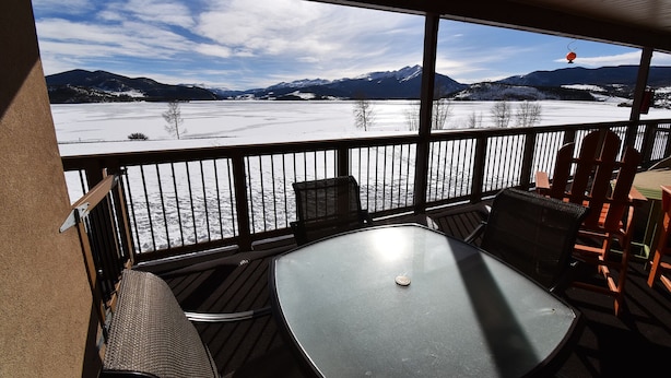 A covered outdoor seating area with a table and chairs, overlooking a frozen lake and mountains. - On Lake! Deck, Views, Elevator, Hot Tub! Walk to Amphitheater, Marina, Dining (Dillon)