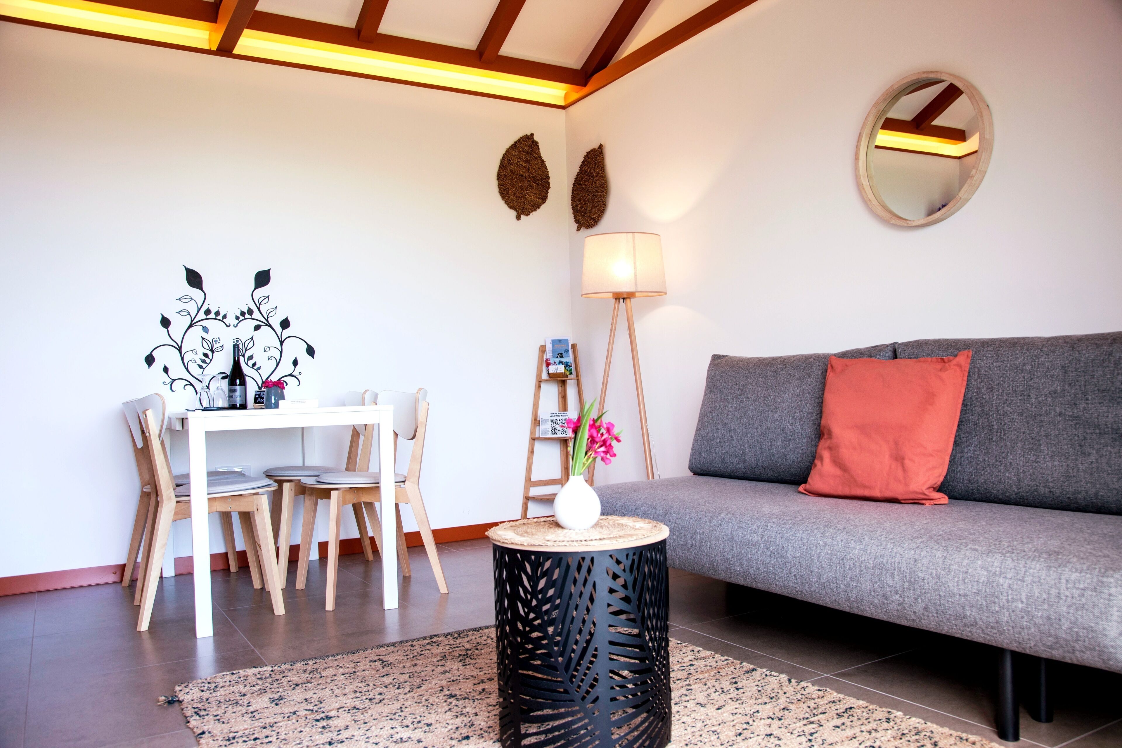 A modern living room with a grey sofa, a round mirror, a dining table with chairs, and a small side table with a vase.