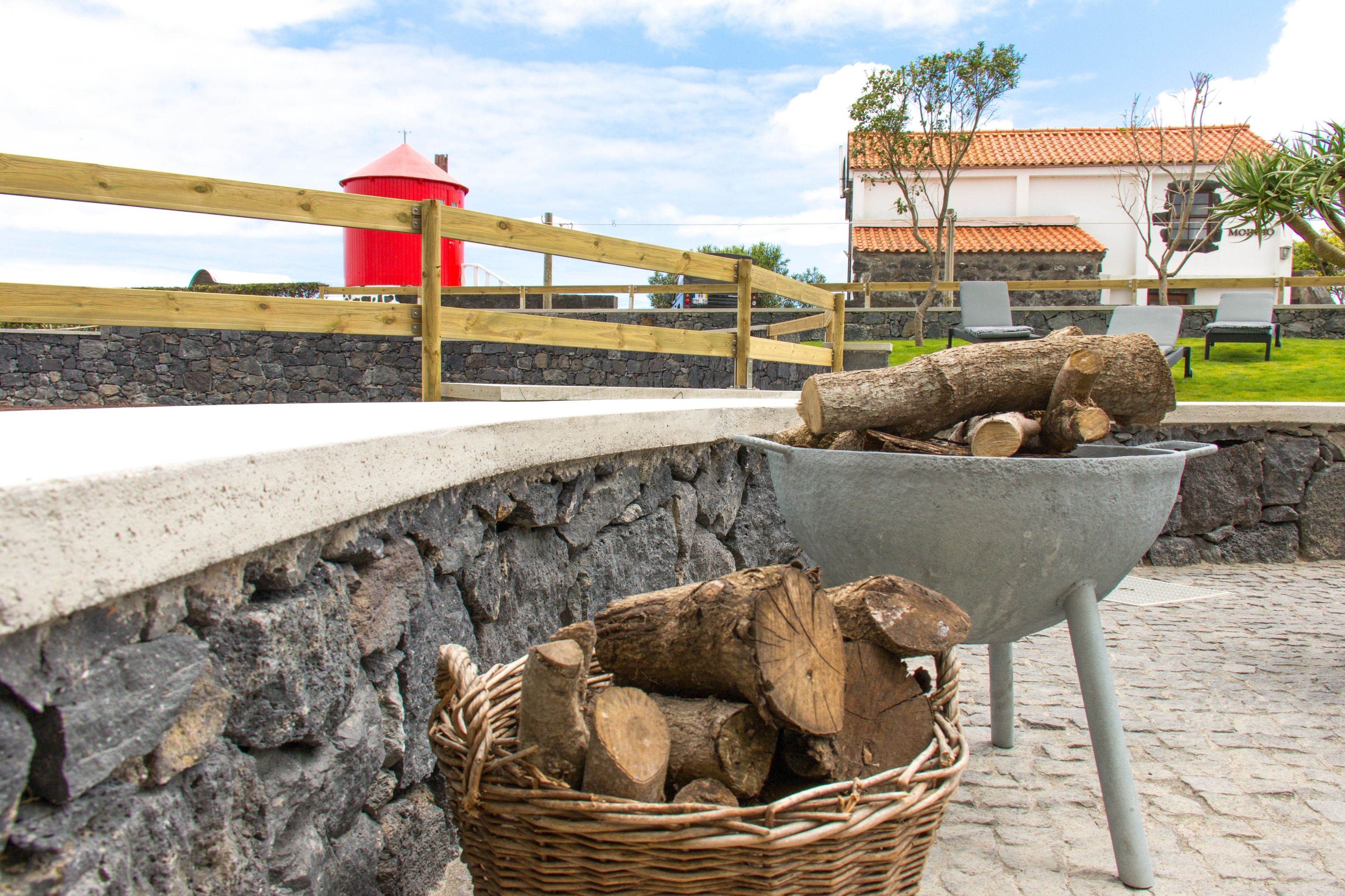 Un muro de piedra con una cesta de mimbre llena de leña, una carretilla con troncos y una estructura cilíndrica roja al fondo.