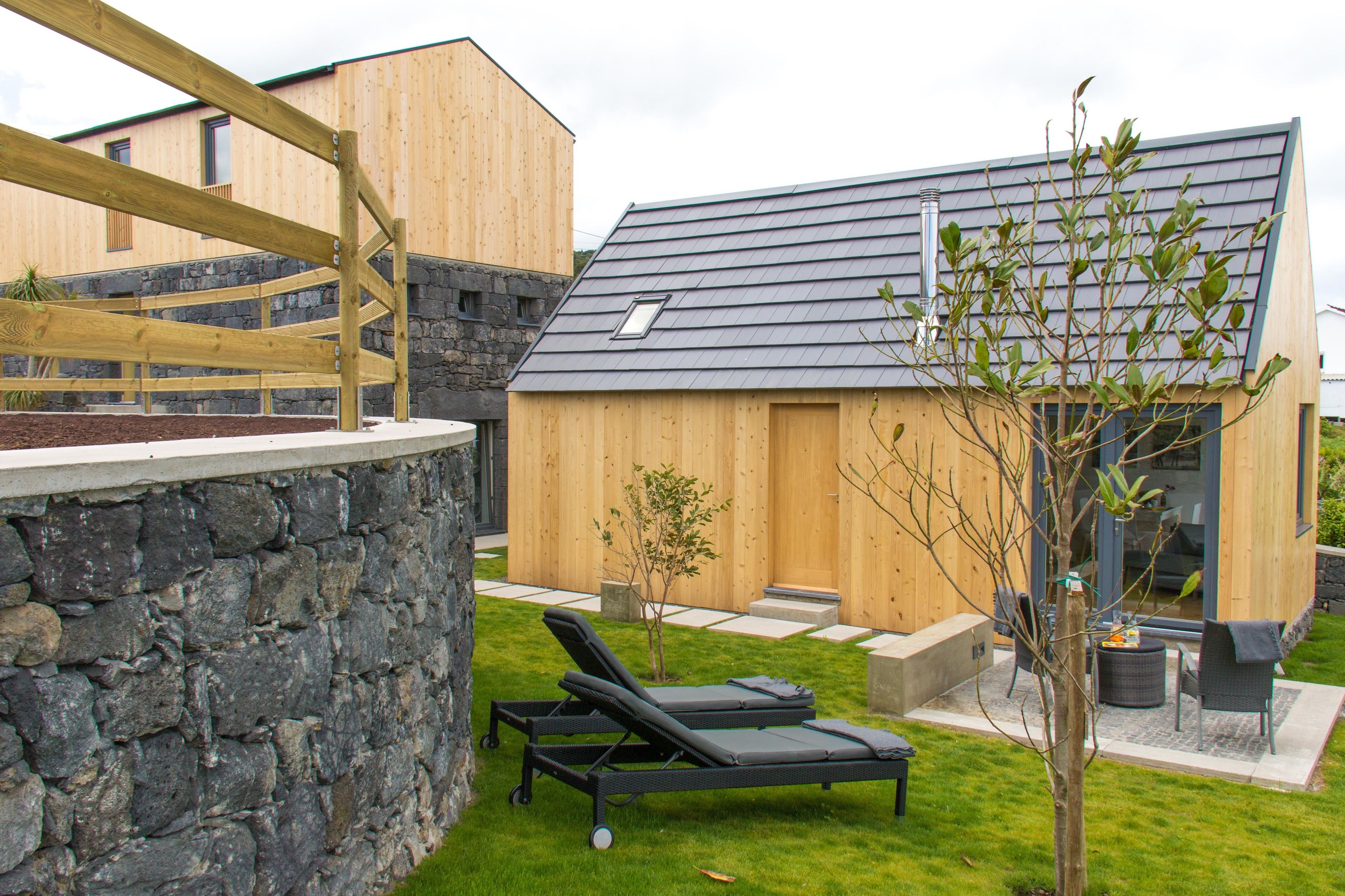 Un espace de détente extérieur moderne, comprenant deux chaises longues, un petit arbre et un bâtiment en bois doté d’un toit en pente.