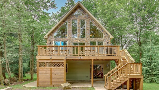 A two-story cabin with a balcony, wooden deck, and a staircase leading to the upper level. - Acorn by PMR, 6 BR Mountain Lodge, Lake View, Hot Tub, Game Room, Fire Pit (Lake Harmony)