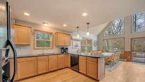 A modern kitchen with wooden cabinets, a dishwasher, and a dining area with chairs. - Acorn by PMR, 6 BR Mountain Lodge, Lake View, Hot Tub, Game Room, Fire Pit (Lake Harmony)