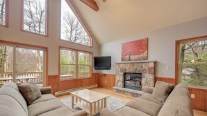 A spacious living room with large windows, a stone fireplace, and a flat-screen TV. - Acorn by PMR, 6 BR Mountain Lodge, Lake View, Hot Tub, Game Room, Fire Pit (Lake Harmony)