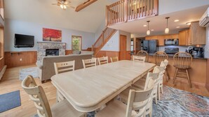 A spacious living area with a large wooden dining table, a cozy fireplace, and a staircase leading to an upper level. - Acorn by PMR, 6 BR Mountain Lodge, Lake View, Hot Tub, Game Room, Fire Pit (Lake Harmony)