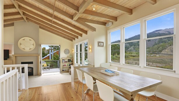 A spacious dining area with a wooden table and white chairs, large windows with a view of the countryside, and a staircase leading to an outdoor area. - Immaculate Beach House with Great Views Of Mt Tam (Stinson Beach)