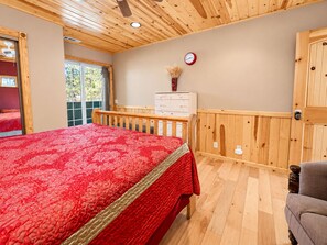 A bedroom with a wooden ceiling, a bed with a red bedspread, a dresser, a clock on the wall, and a window with a view.