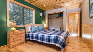 Une chambre avec un plafond en bois, un lit recouvert d’une couverture à carreaux, une commode, une fenêtre avec des stores et une armoire en bois.