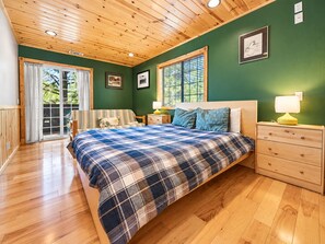 A bedroom with a wooden ceiling, a bed with a plaid bedspread, a nightstand with a lamp, and a window with blinds.