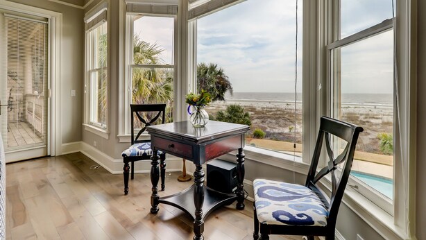 A corner room with large windows overlooking a beach, a table with chairs, and a small plant on the windowsill.