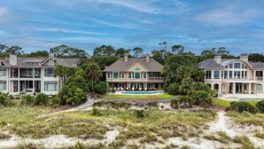 A large, two-story house with a swimming pool in the center, surrounded by lush greenery and a sandy landscape.