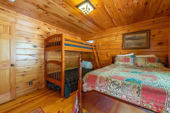 A wooden cabin room with a bunk bed, a single bed with a colorful quilt, and a painting on the wall.