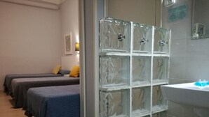 Habitación de hotel con tres camas, una pared de vidrio y un baño con lavamanos y espejo.