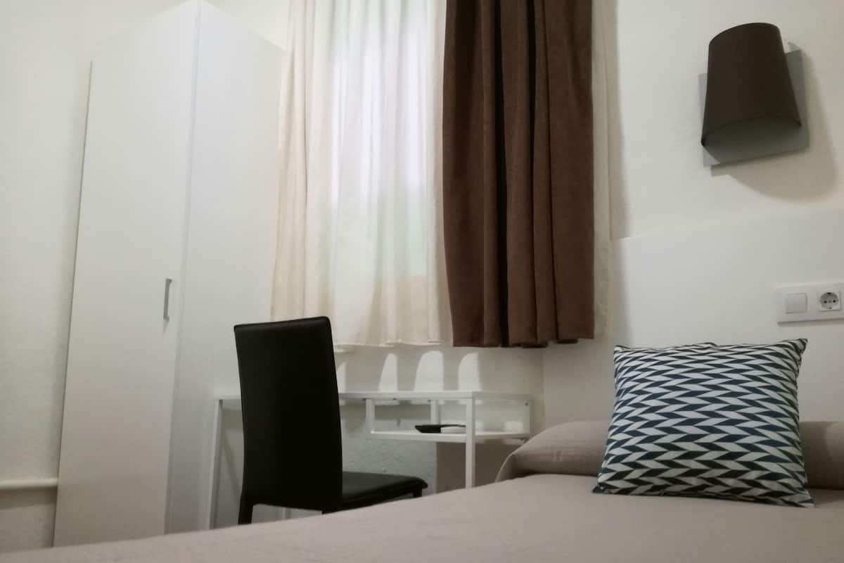 Una habitación con una cama, una silla, un escritorio, una ventana con cortinas y una lámpara fijada a la pared.