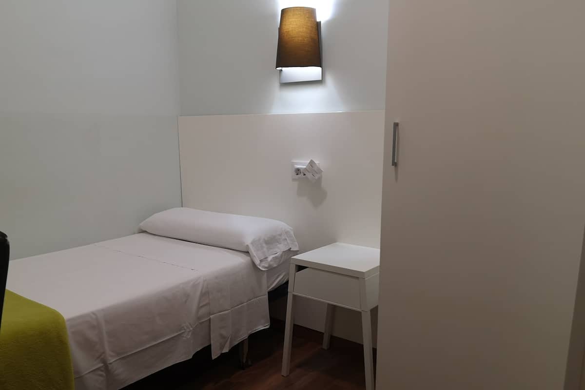 Habitación pequeña y ordenada con una cama individual, un escritorio y una lámpara de pared.