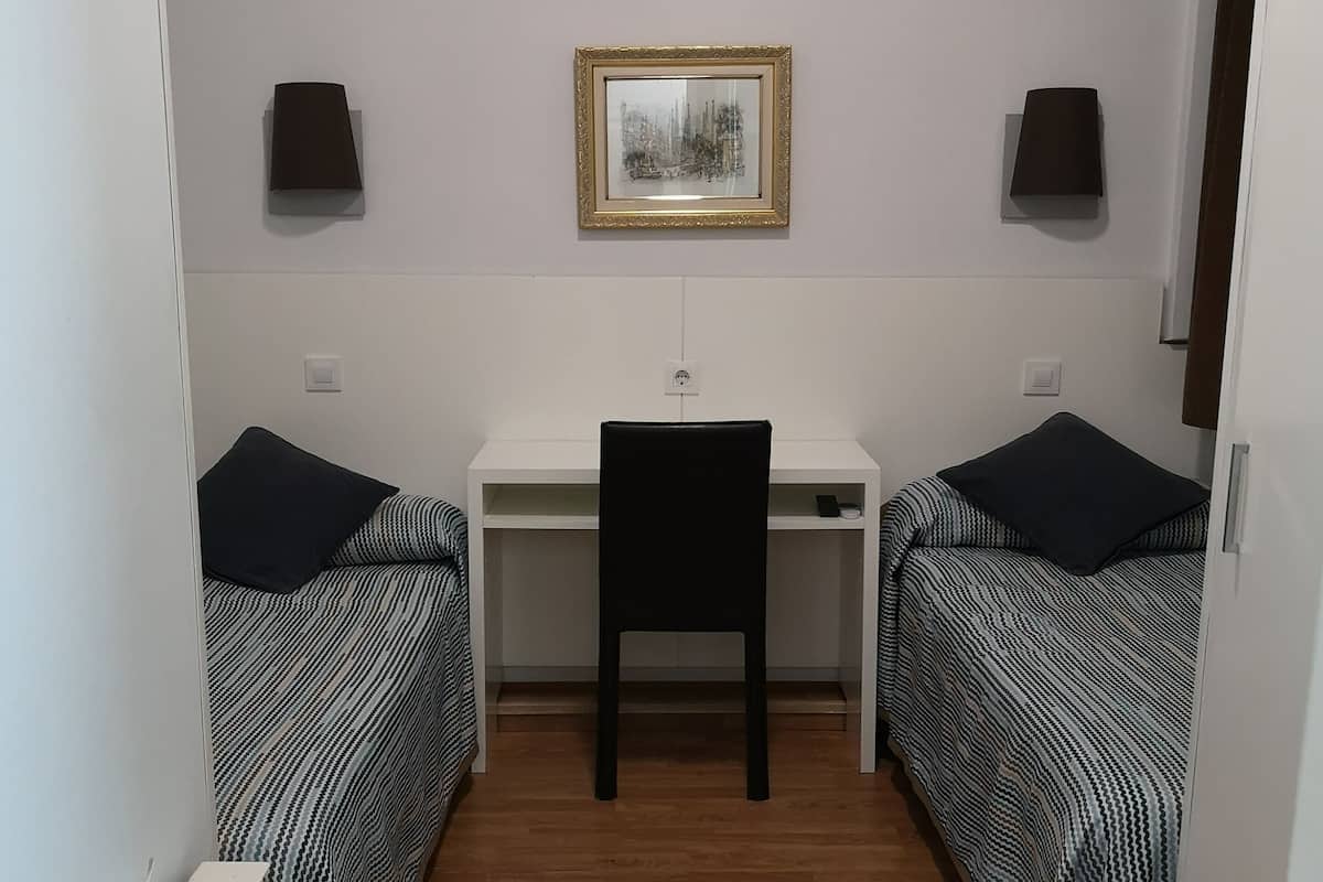 Una habitación pequeña y funcional con dos camas individuales, un escritorio y una silla. Hay dos lámparas de pared y un cuadro enmarcado sobre el escritorio.