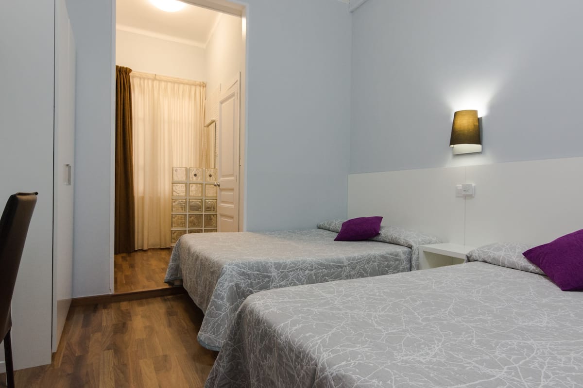 Una habitación con dos camas, una mesita de noche, una lámpara de pared y una puerta que da a otra estancia.