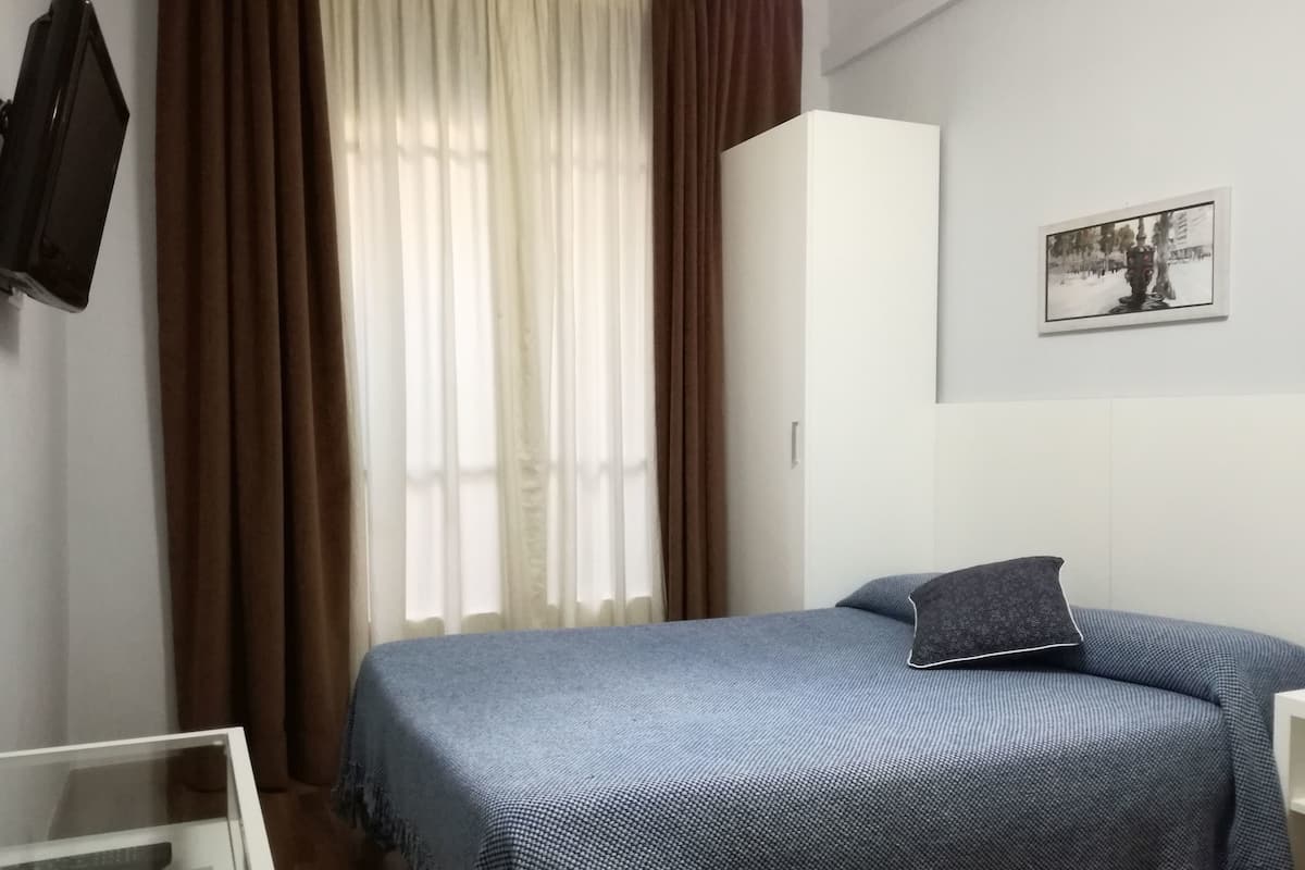 Un dormitorio con cama, armario, televisor y ventana con cortinas.
