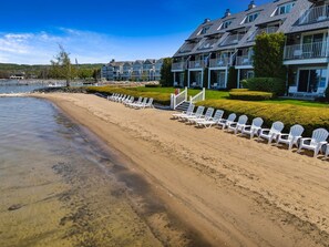 Un hotel frente a la playa con varios balcones, una vista despejada del agua y una fila de sillas de playa blancas a lo largo de la orilla.