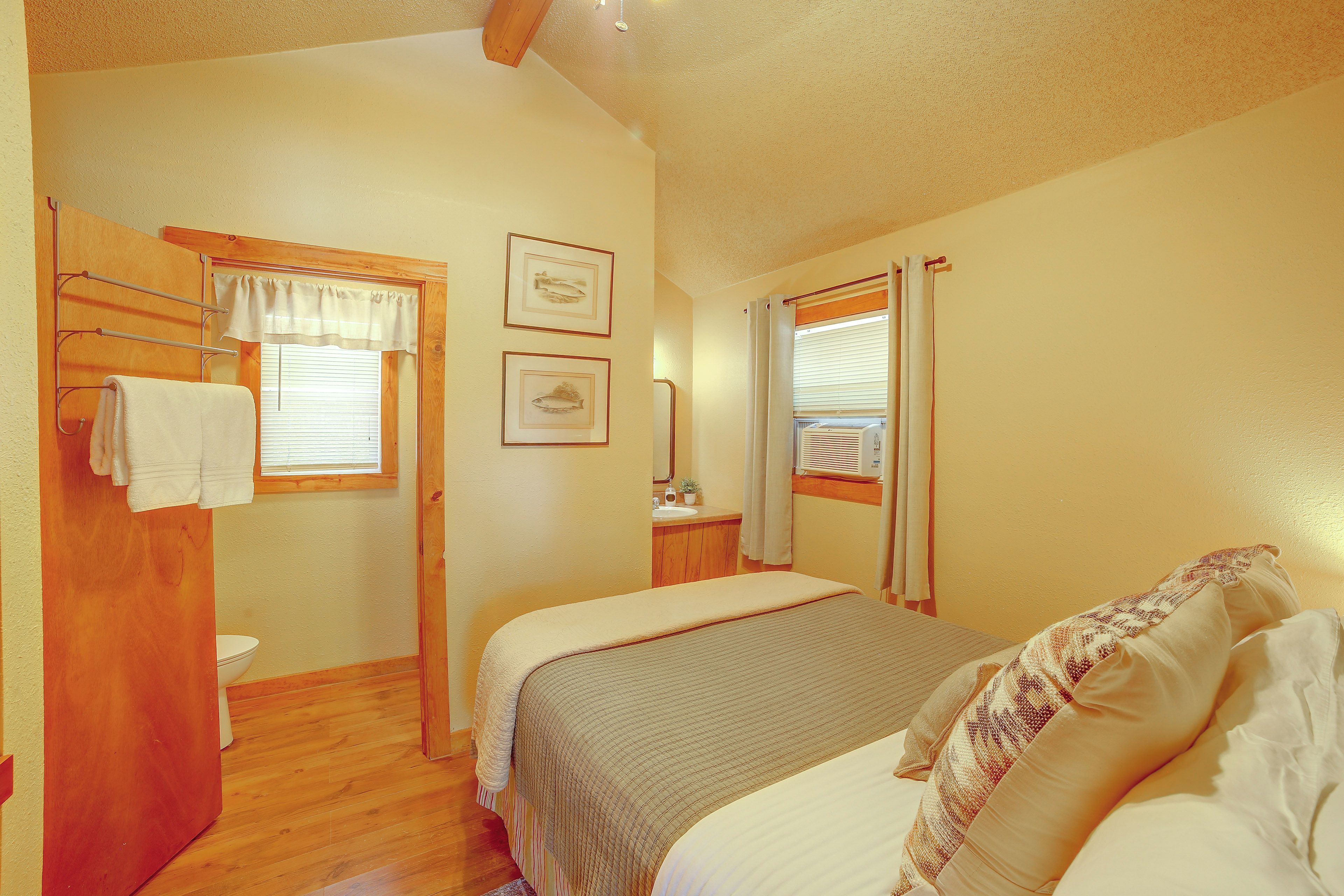 A bedroom with two beds, a wooden floor, a bathroom with a toilet and sink, and a window with blinds.