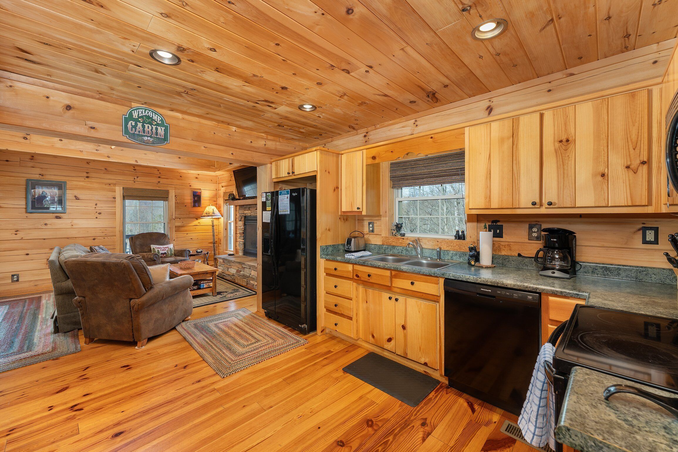 A cabin interior with wooden walls and floors, a sofa, a dining table, a kitchen with a refrigerator, stove, and sink.