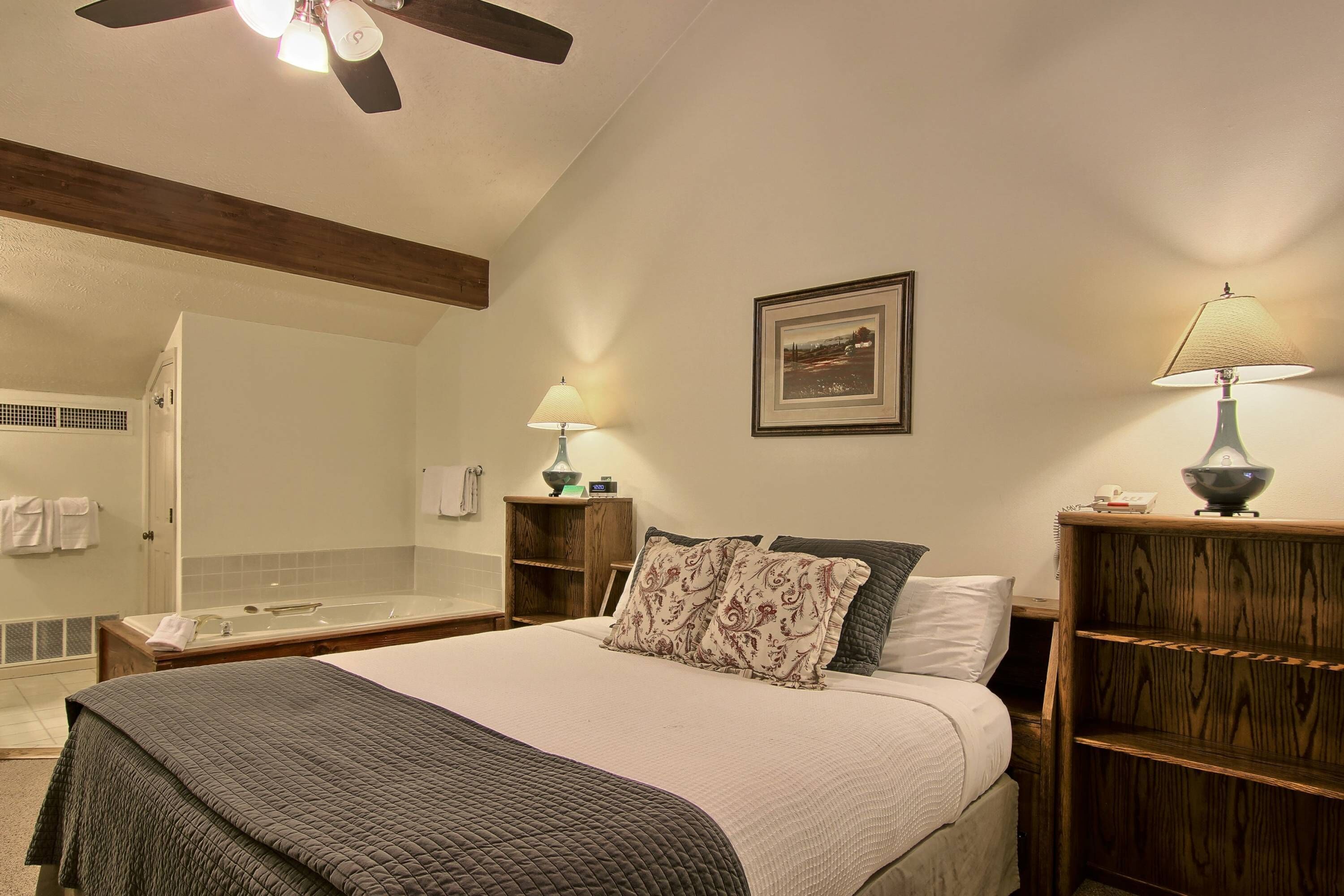 Hotelkamer met een groot bed, een houten nachtkastje, een badkamer met een glazen douche en een plafondventilator.
