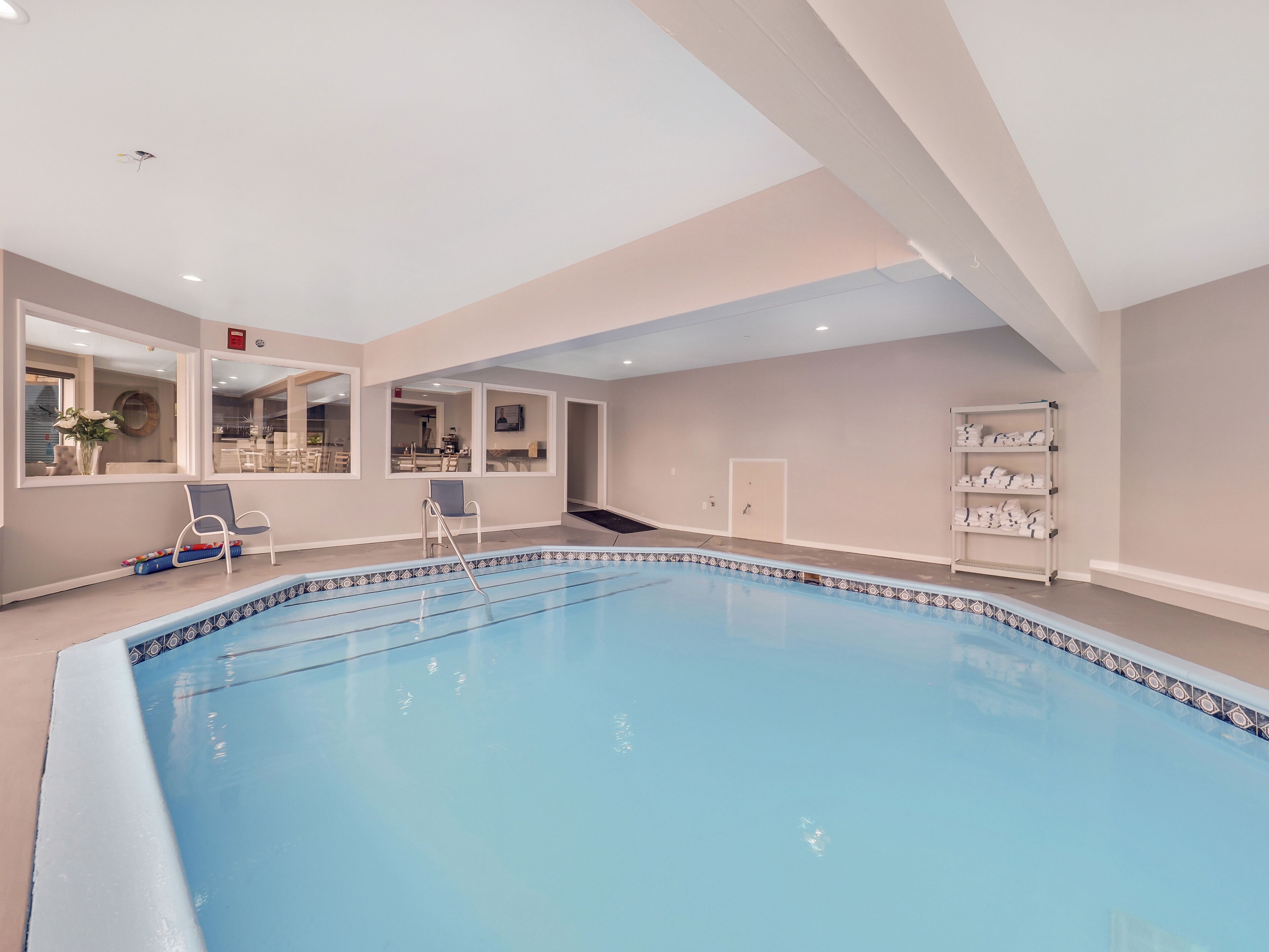 An indoor swimming pool with a clear view of the adjacent dining area, a chair by the pool, and a shelf with towels.