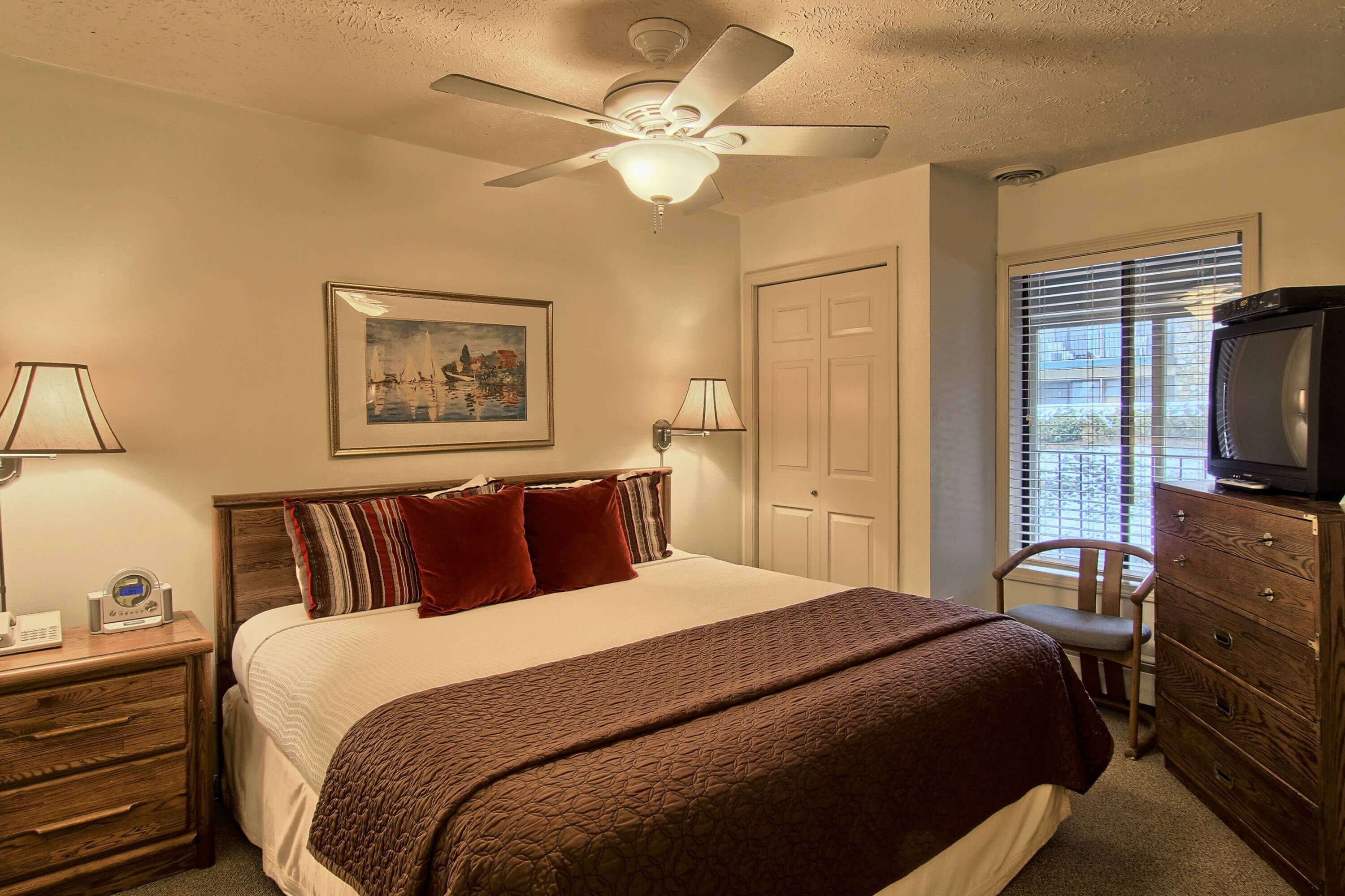 A hotel room with a bed, a ceiling fan, a television, a dresser, a chair, and a window with blinds.