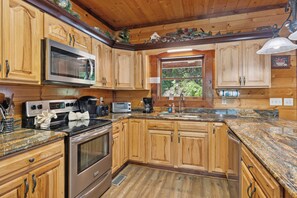 A kitchen with wooden cabinets, a microwave, a stove, a sink, and a window with a view of greenery.