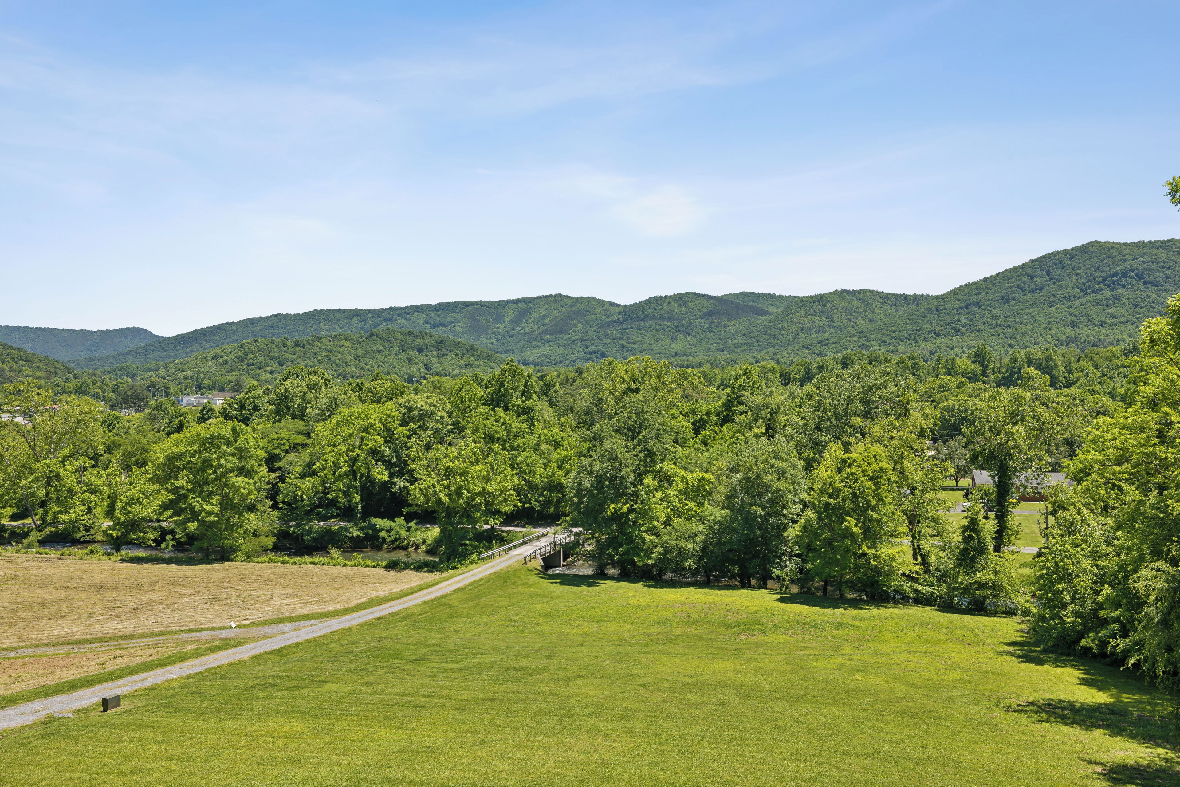 A grassy field with a pathway leading to a bridge over a stream, surrounded by dense forest and hills.