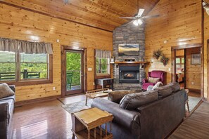 Interior de una cabaña de madera con chimenea de piedra, sofá de cuero y vistas al exterior a través de amplios ventanales.