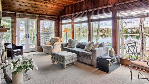 Una sala de estar acogedora con techo de madera, ventanas amplias, un sofá gris, un sillón de cuero negro y una mesita con una lámpara.