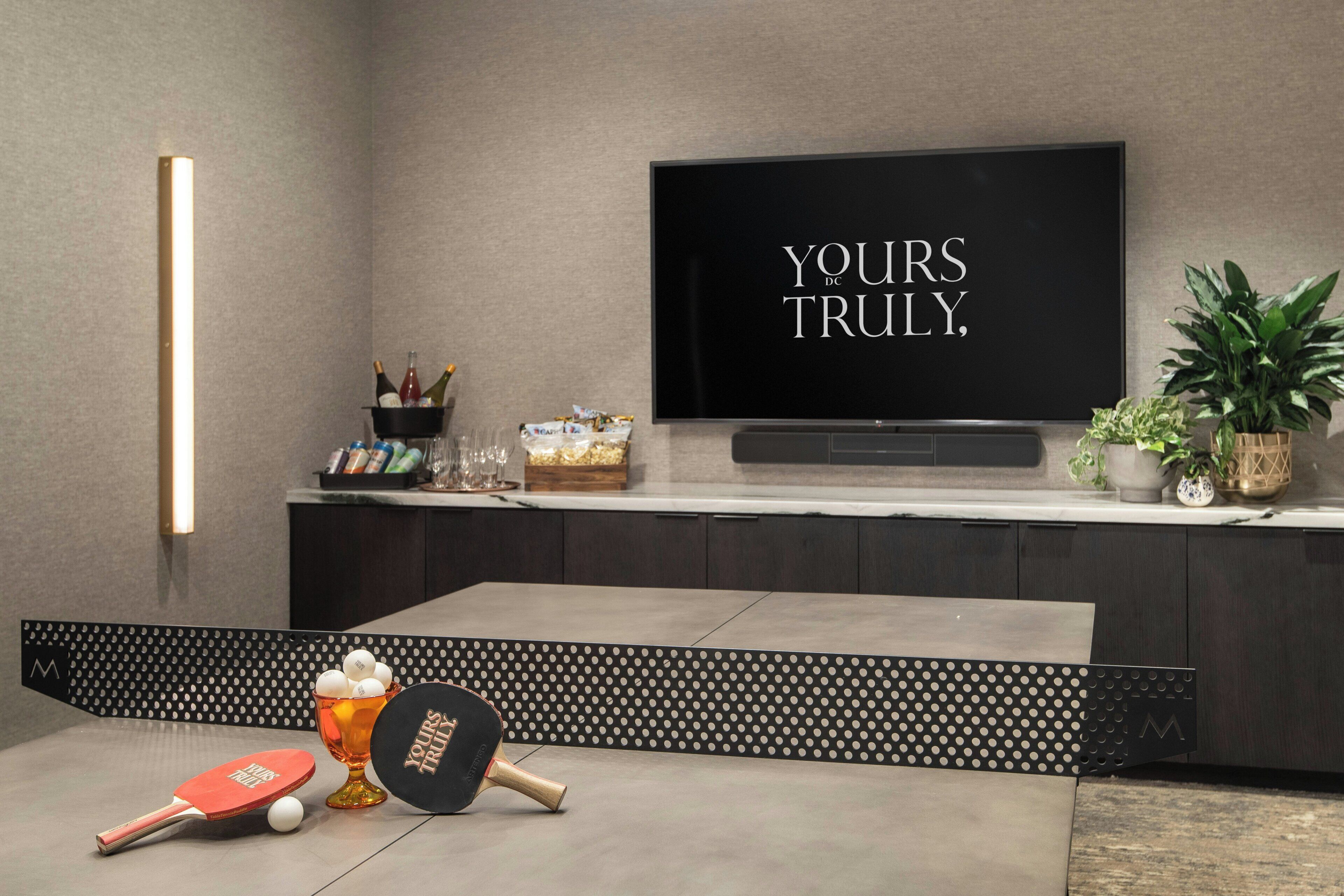 A modern living room with a flat-screen TV mounted on the wall, a ping pong table, and a glass of beer.