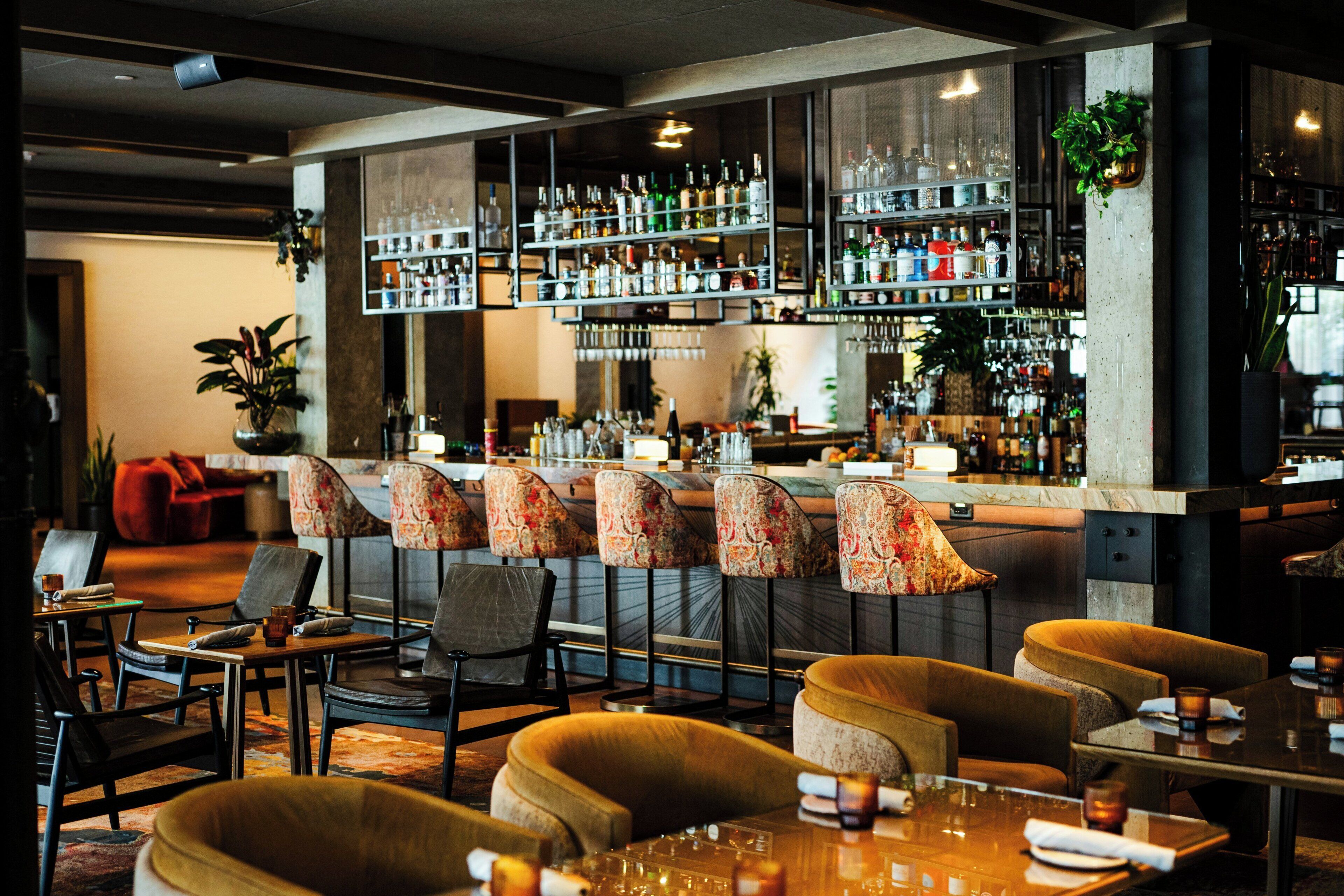 A bar area with multiple seating options, including bar stools and cushioned chairs, with a variety of bottles and glasses on shelves.