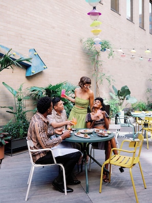 A group of people dining outdoors at a table with chairs, surrounded by potted plants and a brick wall.