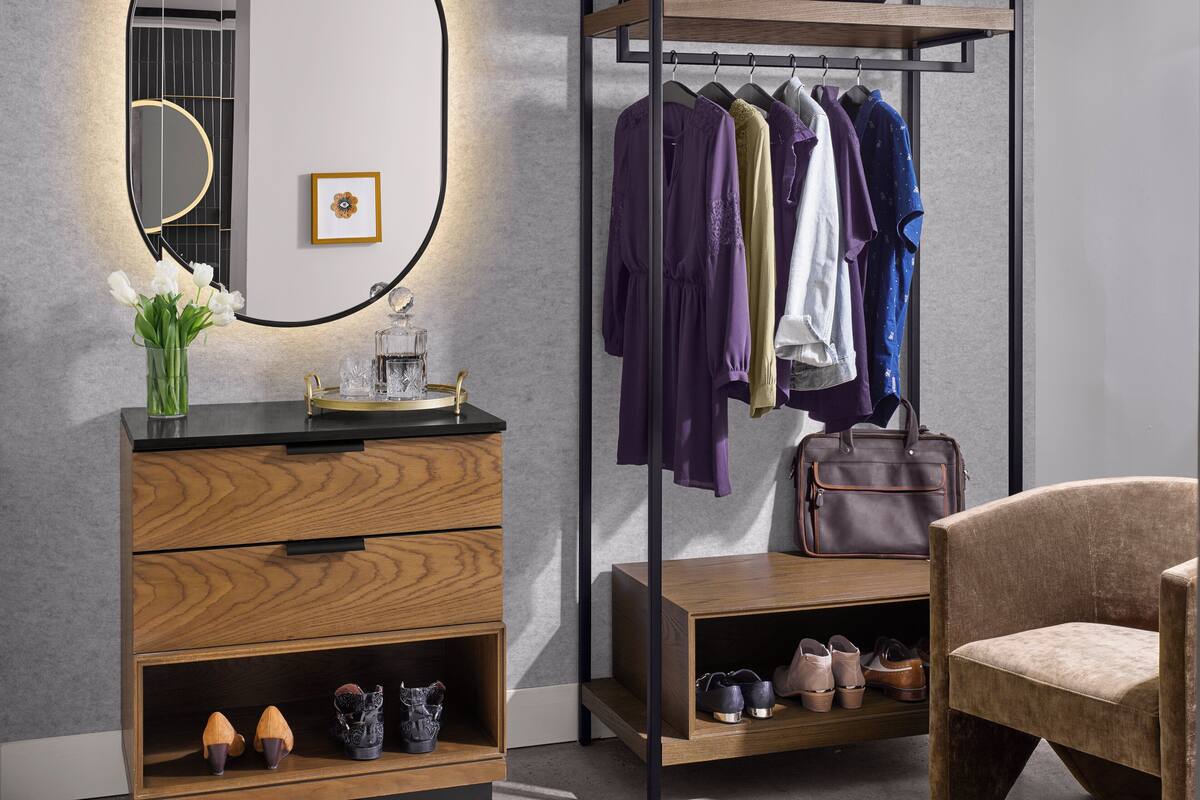 A modern room with a wooden dresser, a mirror, a coat rack with clothes, a suitcase, and a chair.
