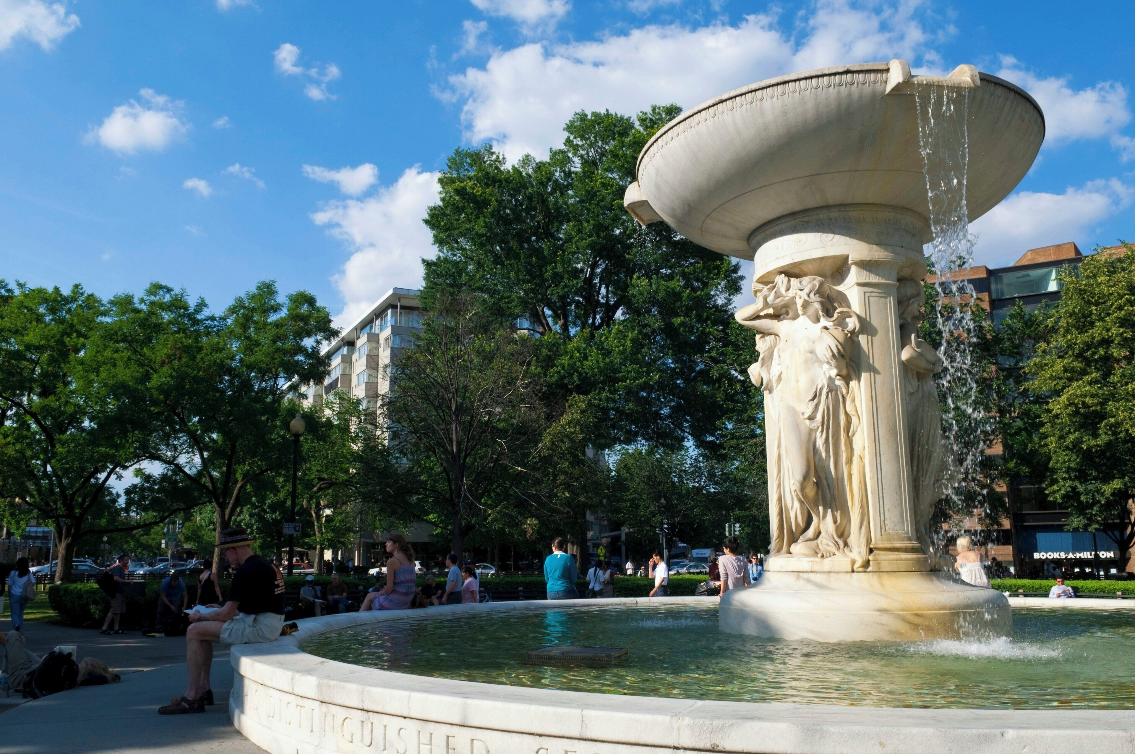 Ein Brunnen mit großem Becken und Statuen, umgeben von Bäumen und Personen.