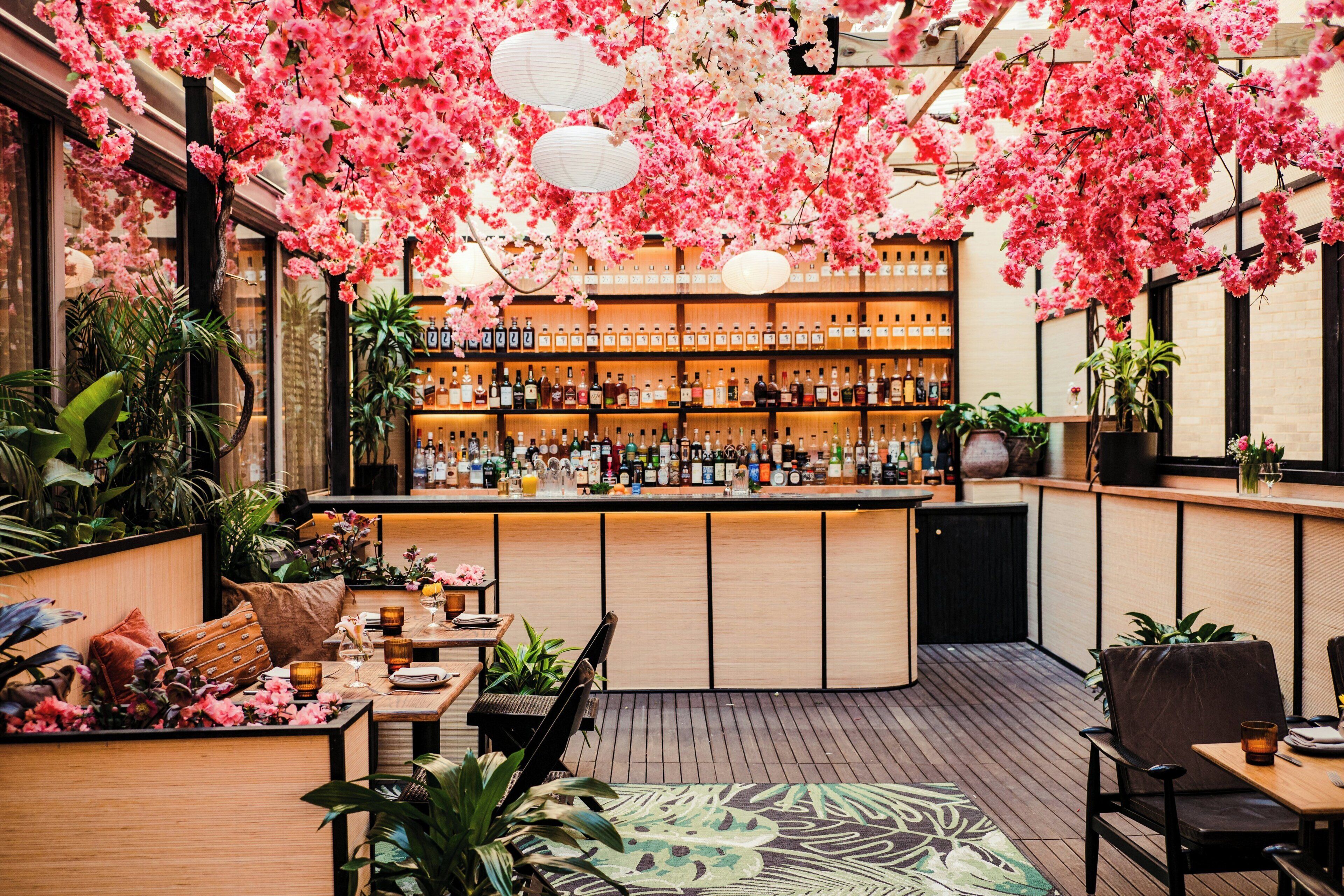 A bar area with a wooden counter, shelves of bottles, and hanging pink flowers.