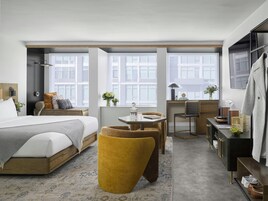 A modern hotel room with a bed, a small table, a chair, a desk, and a wardrobe.