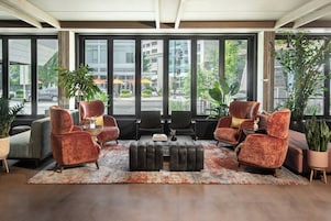 A spacious lobby with large windows, leather armchairs, a central ottoman, and potted plants.