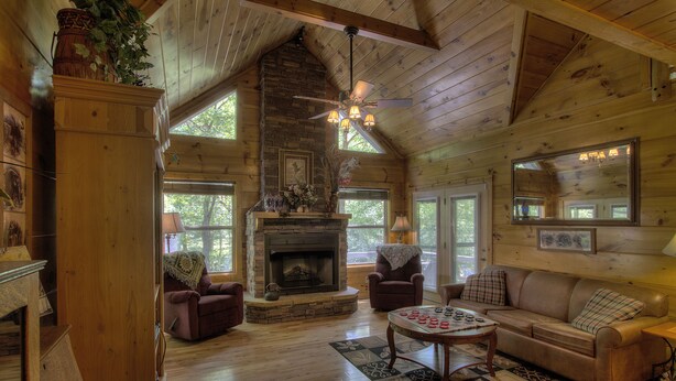 A cozy cabin interior with a stone fireplace, wooden walls, a ceiling fan, and a large window offering a view of the outdoors.