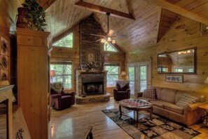 A cozy cabin interior with a stone fireplace, wooden walls, a ceiling fan, and a large window offering a view of the outdoors. - Creekside Cabin-Private Hot Tub-Firepit-Fishing (Cosby)