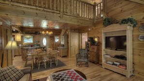 A wooden cabin interior with a dining area, a living room with a television, and a kitchen.