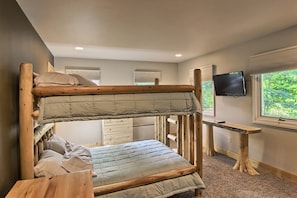 A bunk bed with a wooden frame, a desk with a chair, a television, and a window with a view of trees.