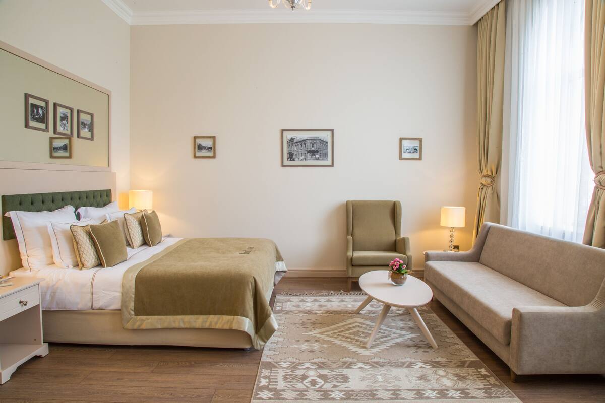 A hotel room with a bed, a sofa, a chair, a small table, and a lamp. There are framed pictures on the wall and a window with curtains.