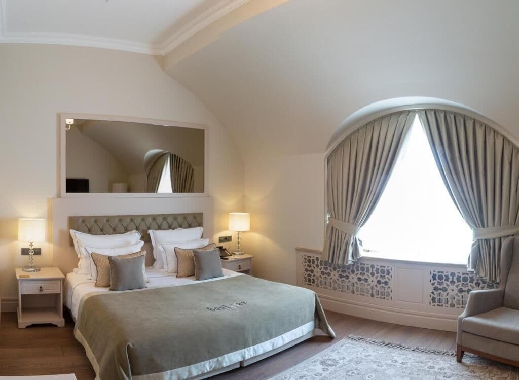 A bedroom with a large bed, two bedside tables, a nightstand, a chair, a window with curtains, and a decorative wall panel.