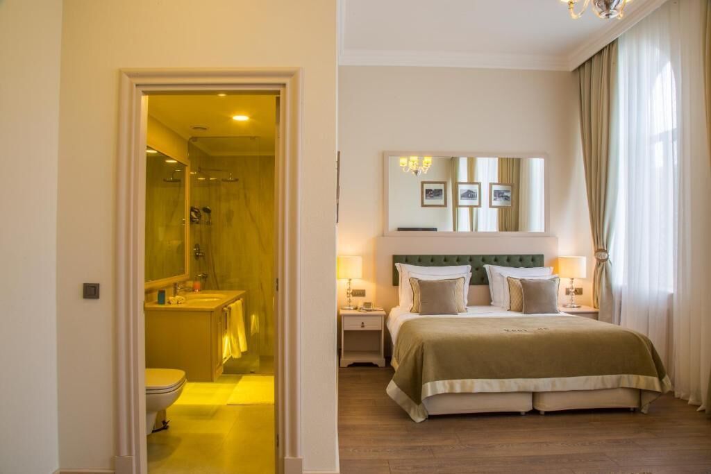 Une chambre d’hôtel avec un lit, une table de chevet, une commode, un miroir et une salle de bain visible à travers une porte ouverte.