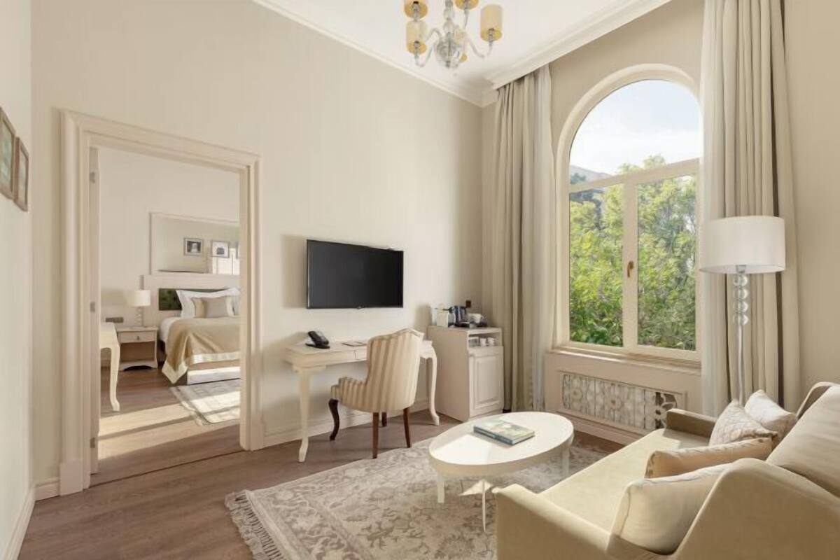 A hotel room with a bed, a desk, a TV, a sofa, and a large window with a view of greenery.