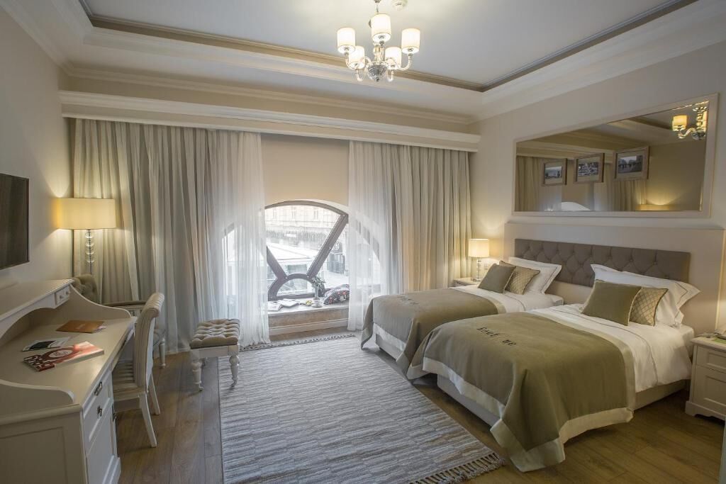 A hotel room with two beds, a desk, a chair, a TV, and a large window with curtains.